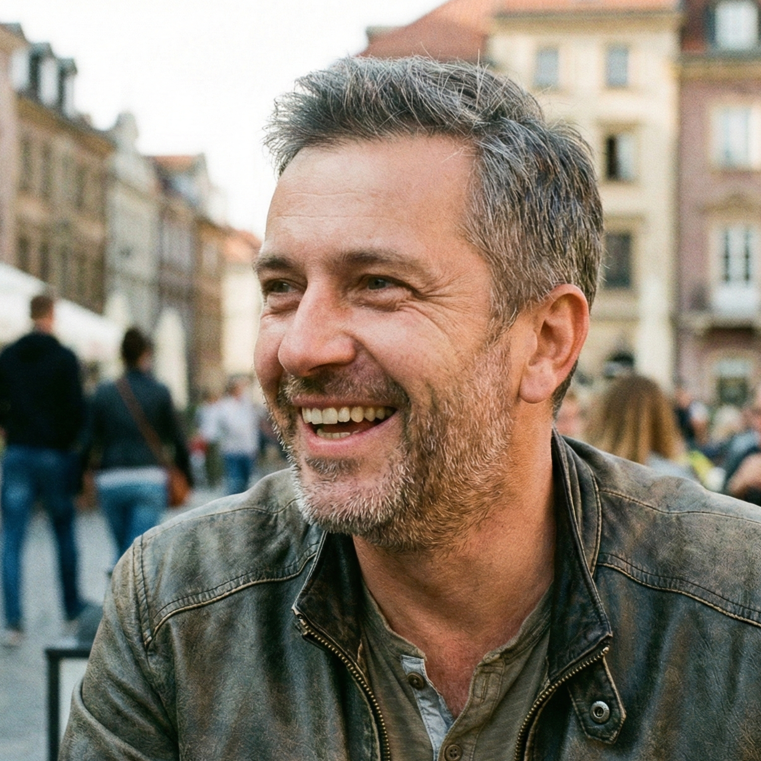 Uśmiechnięty mężczyzna w skórzanej kurtce siedzi w kawiarni na tle miejskiego krajobrazu, ludzie spacerują w tle.