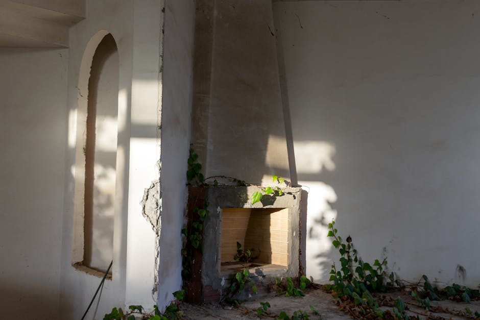 cover-image-276839 Zniszczony kominek w opuszczonym wnętrzu, otoczony zielonymi pnączami. Ściany w jasnym kolorze, z widocznymi cieniami i fragmentami tynku. Idealne miejsce do fotografii architektury i urban exploration.