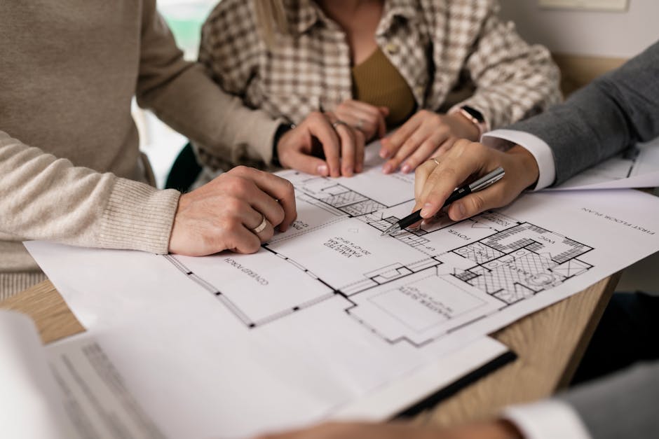 Grupa osób pracująca nad planem architektonicznym, z bliskim ujęciem rąk trzymających rysunki i długopisy. W tle widać elementy biurowe.