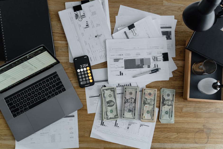 Stół z dokumentami finansowymi, kalkulatorem i gotówką. Widoczne są wykresy i analizy na arkuszach papieru oraz laptop z otwartym programem do zarządzania danymi.